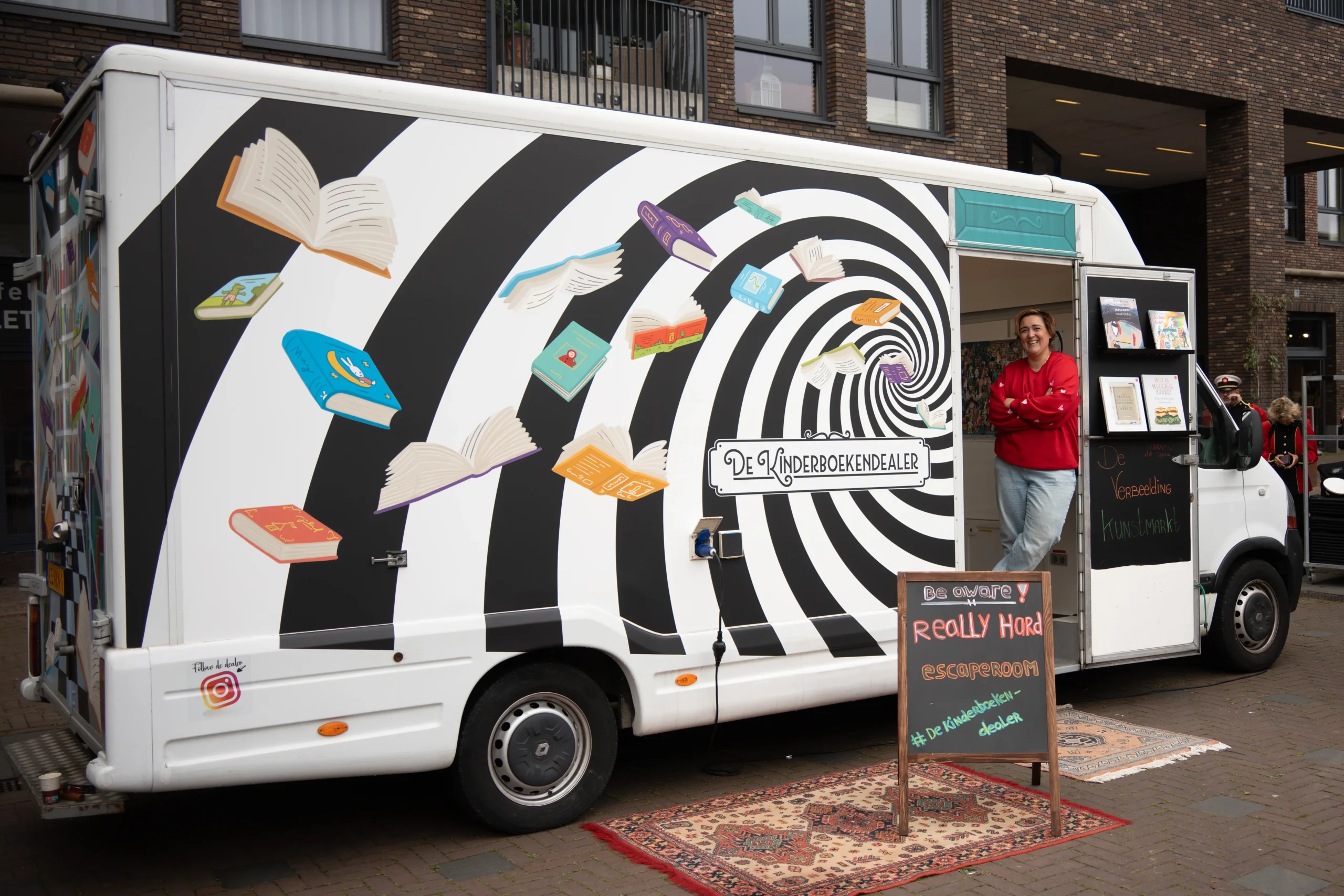 De Kinderboekbus van De Kinderboekendealer staat geparkeerd met een voorlezer in de deuropening en een bord met activiteiten ervoor.