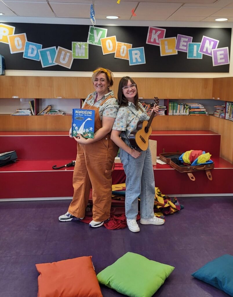 Tamara van De Kinderboekendealer en een collega geven een muzikale voorleesvoorstelling met boek en ukelele in de bibliotheek.
