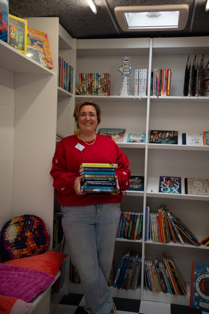 Tamara De Kinderboekendealer in de boekenbus met een stapel kinderboeken – rijdende kinderboekenwinkel en voorleesvoorstellingen.