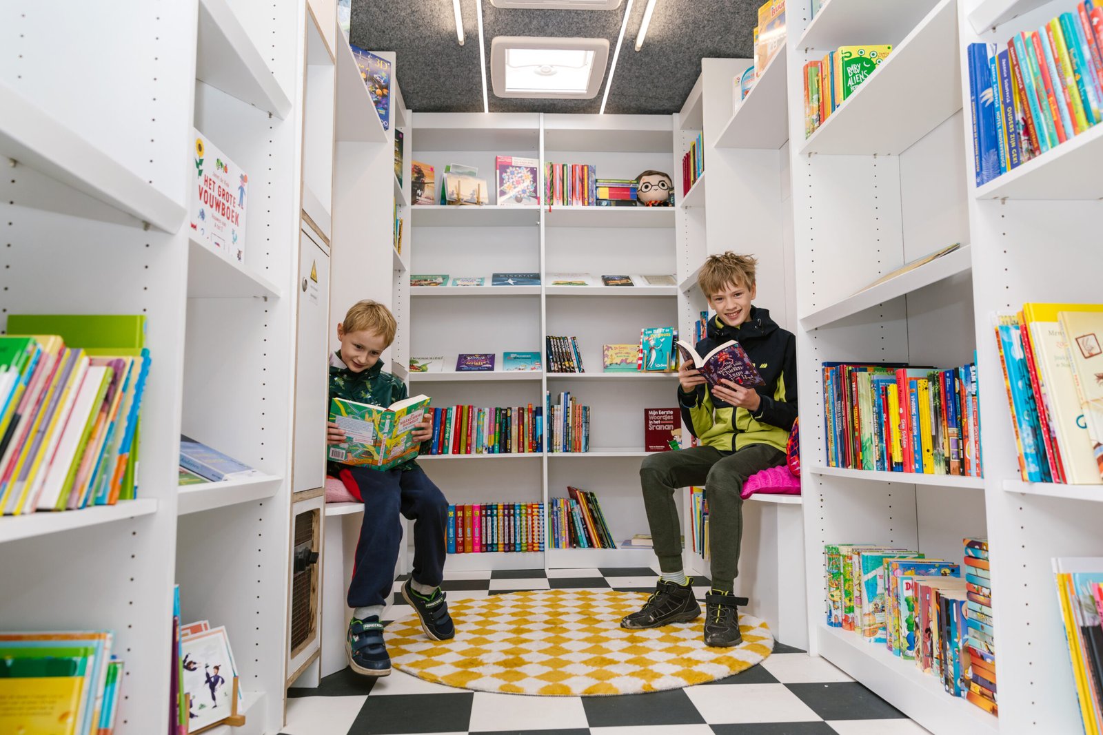 Twee kinderen lezen in de boekenbus van De Kinderboekendealer, een rijdende kinderboekenwinkel vol leesplezier voor scholen en bibliotheken.